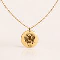 A gold chain necklace with a round engraved pendant featuring a dog portrait and the name 'Rudy' at the bottom, displayed on a light cream background with a soft reflection.