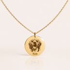 A gold chain necklace with a round engraved pendant featuring a dog portrait and the name 'Rudy' at the bottom, displayed on a light cream background with a soft reflection.